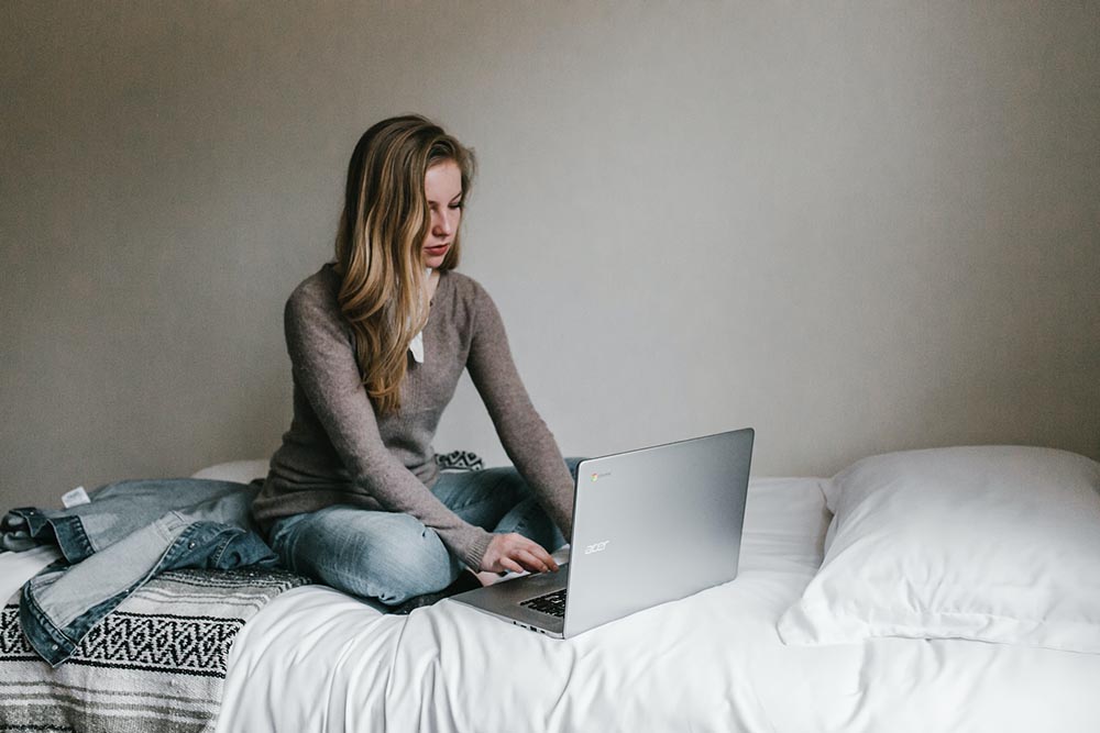 woman sat cross-legged on a bed using a laptop, with a denim jacket folded next to her.