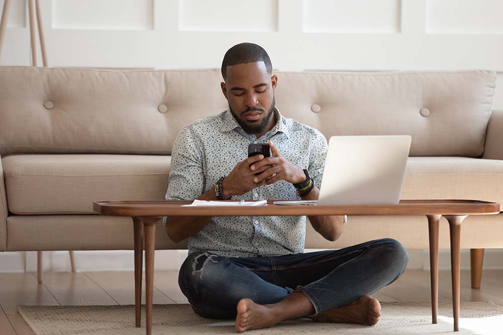 man browsing the internet using a smartphone and a laptop, sat cross-legged on the floor