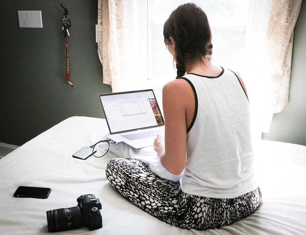 young woman sat cross-legged on a bed using a laptop, with a digital camera and smartphone beside her.